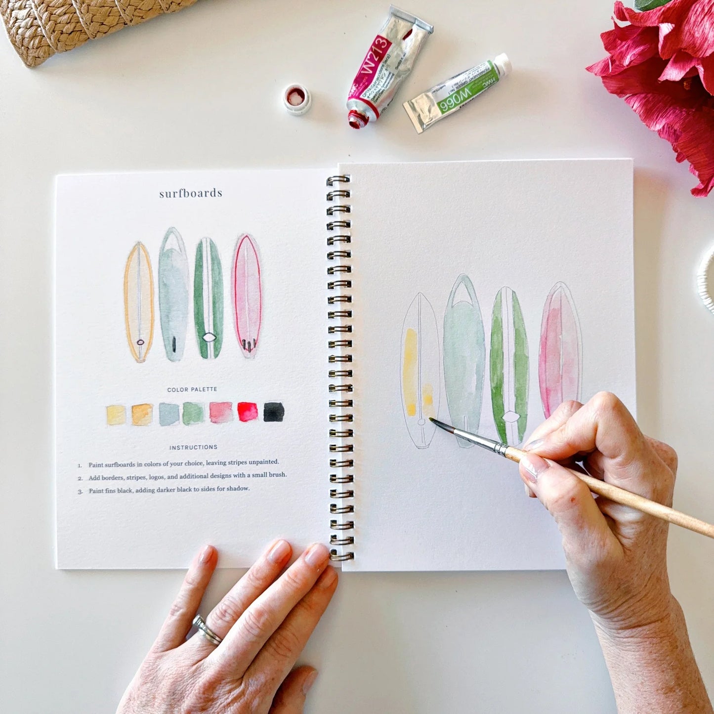 Person coloring surfboard illustrations in a coloring book with art supplies on a table.