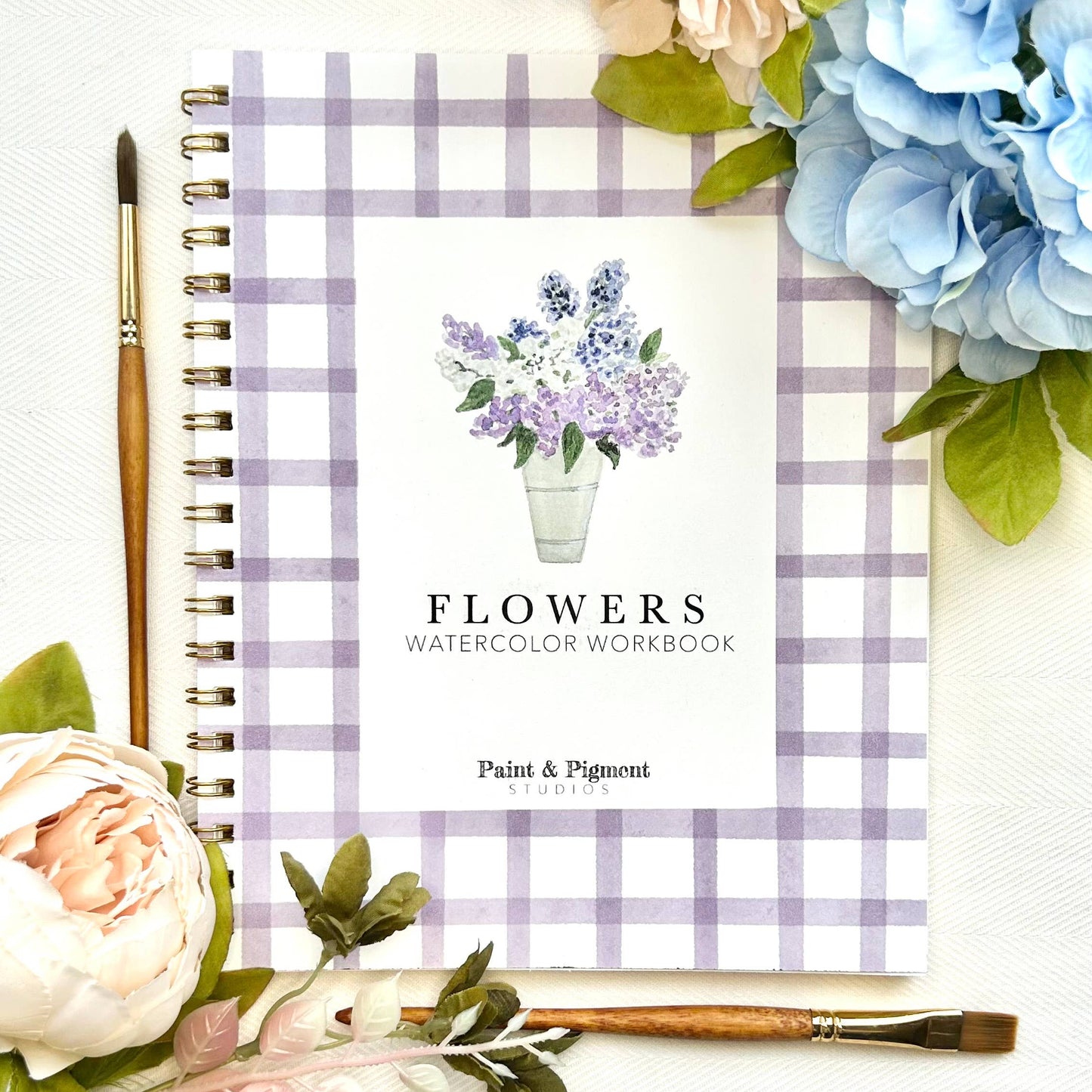 Flowers watercolor workbook with plaid cover, surrounded by flowers and a paintbrush on a white background