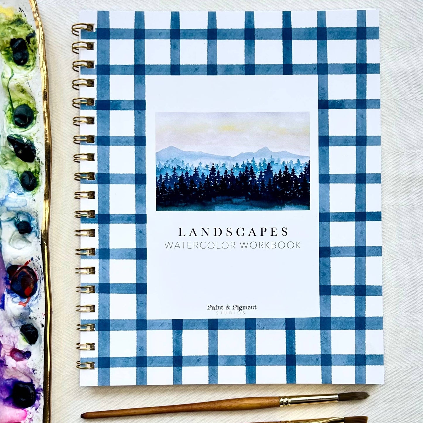 Workbook titled 'Landscapes Watercolor Workbook' with blue plaid cover, paintbrushes, and art supplies on a light background