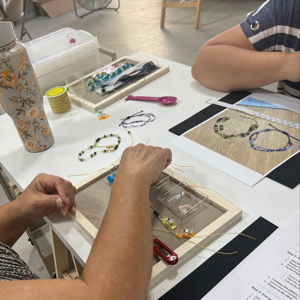A close-up of a person's hands working at a craft table, assembling a beaded jewelry piece in a wooden frame. The table is filled with craft supplies, including a floral-patterned water bottle, beads, and printed instruction sheets.