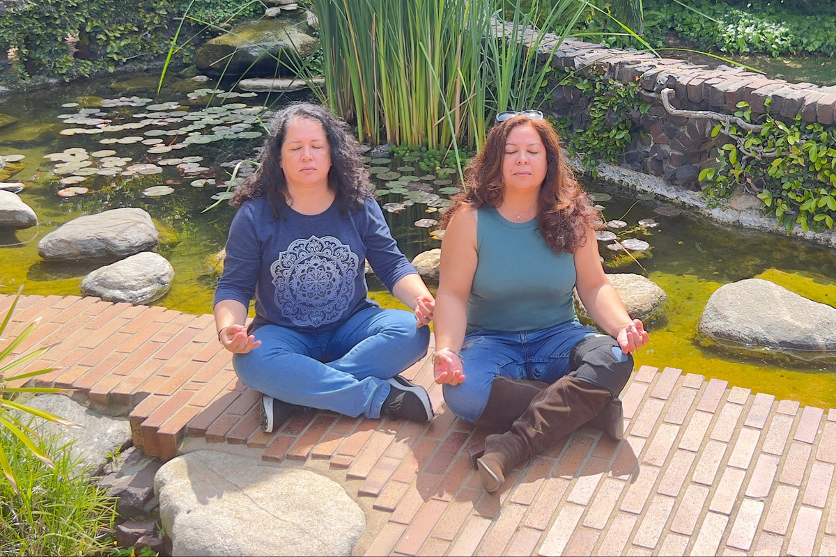 Two women meditating outdoors by a small pond with rocks, green reeds, and a stone wall in a landscaped garden.
