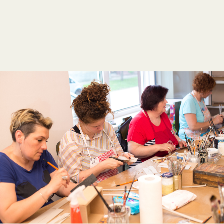 Four women seated at a table in a craft or art studio, working with paintbrushes, paper, and small tools. The scene is indoors with a window in the background and a light beige border around the photo.