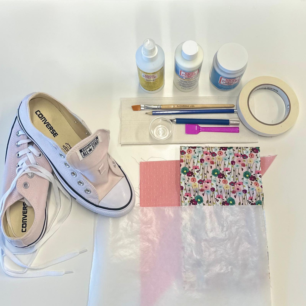 A flat-lay of a DIY sneaker restoration or customization kit on a white surface, featuring a light pink Converse sneaker, acrylic glue, paint, masking tape, and floral fabric pieces.