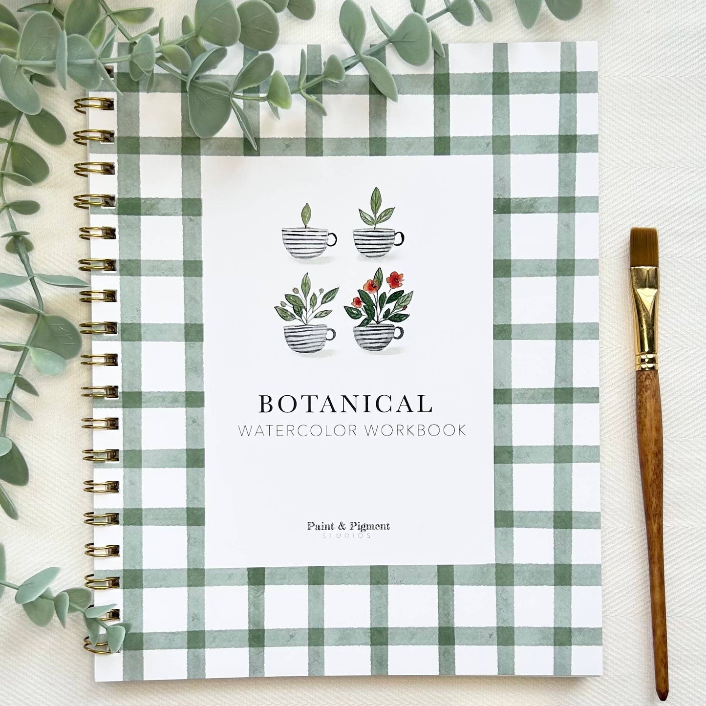 Botanical watercolor workbook with plaid cover, surrounded by eucalyptus leaves and a paintbrush.