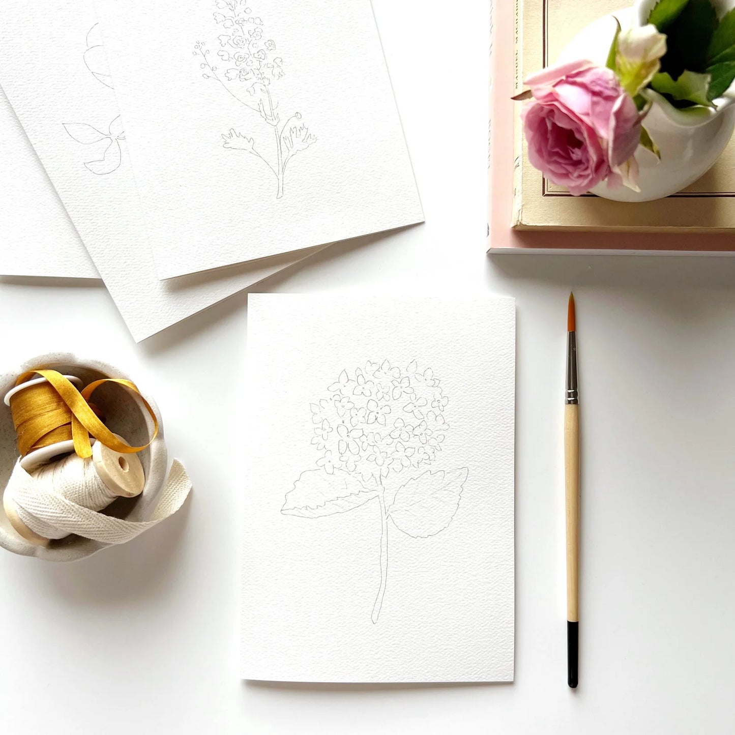 Artistic setup with white paper, paintbrush, spool of thread, and pink rose on a white surface.