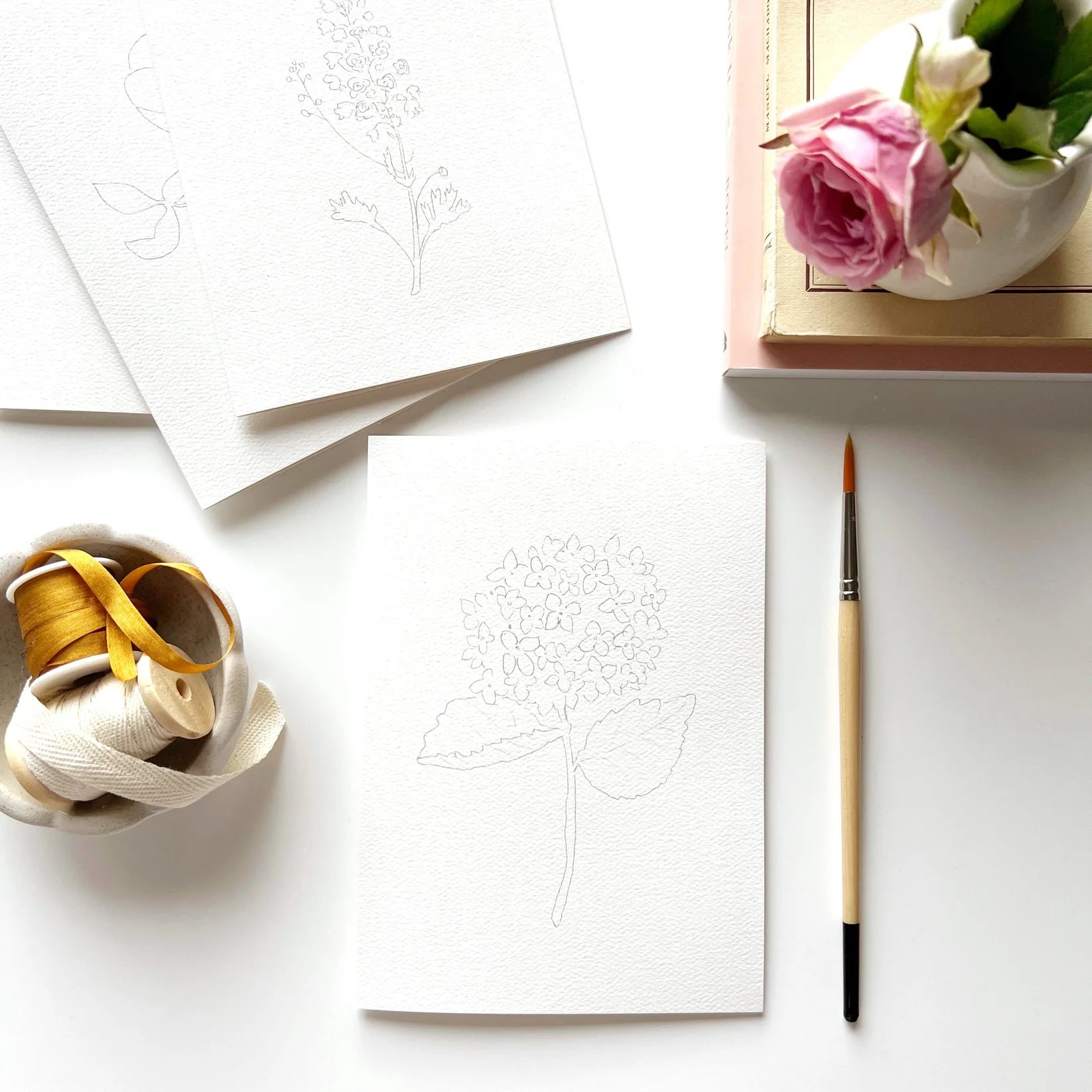 Artistic setup with white paper, paintbrush, spool of thread, and pink rose on a white surface.