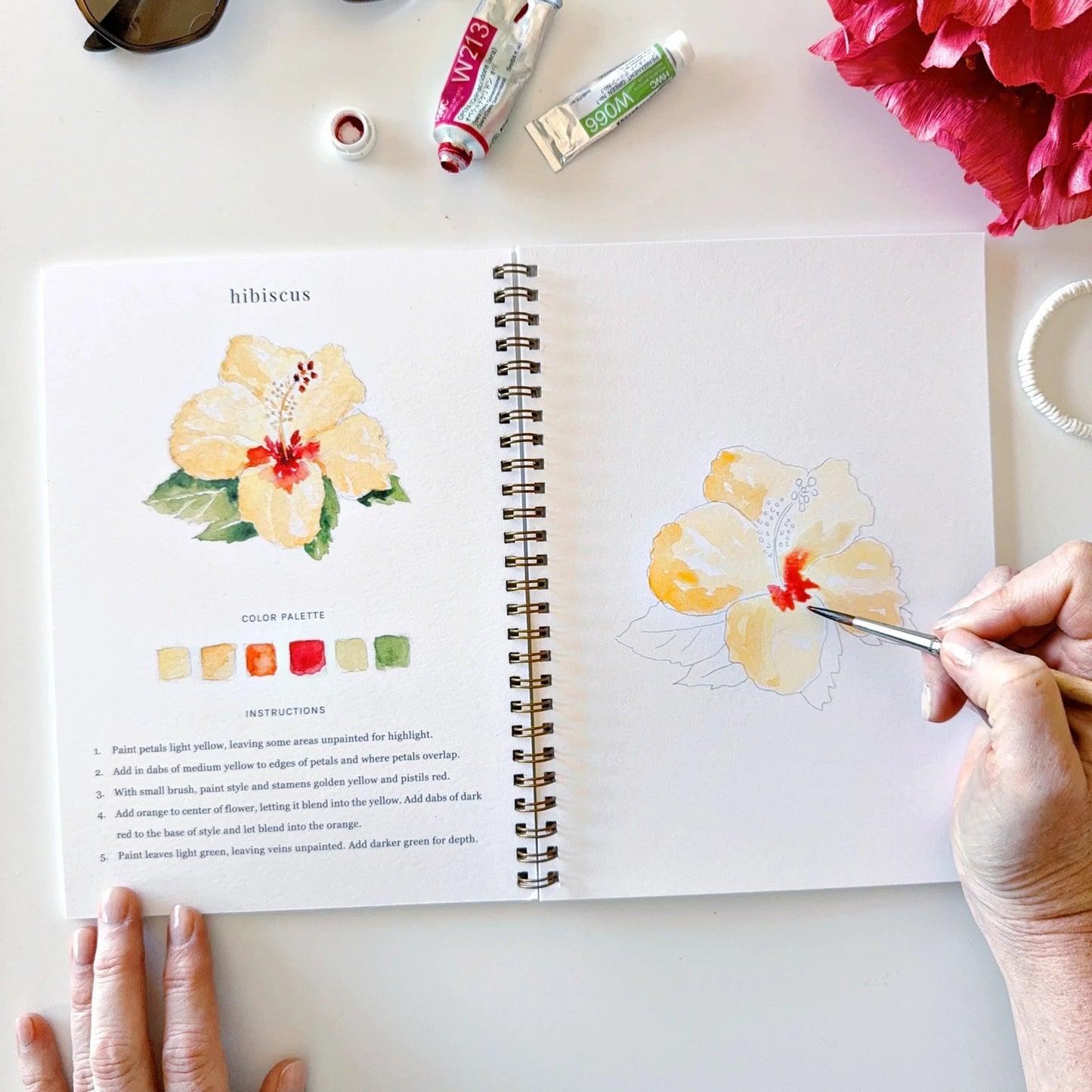 Person painting a hibiscus flower in a sketchbook with art supplies around.
