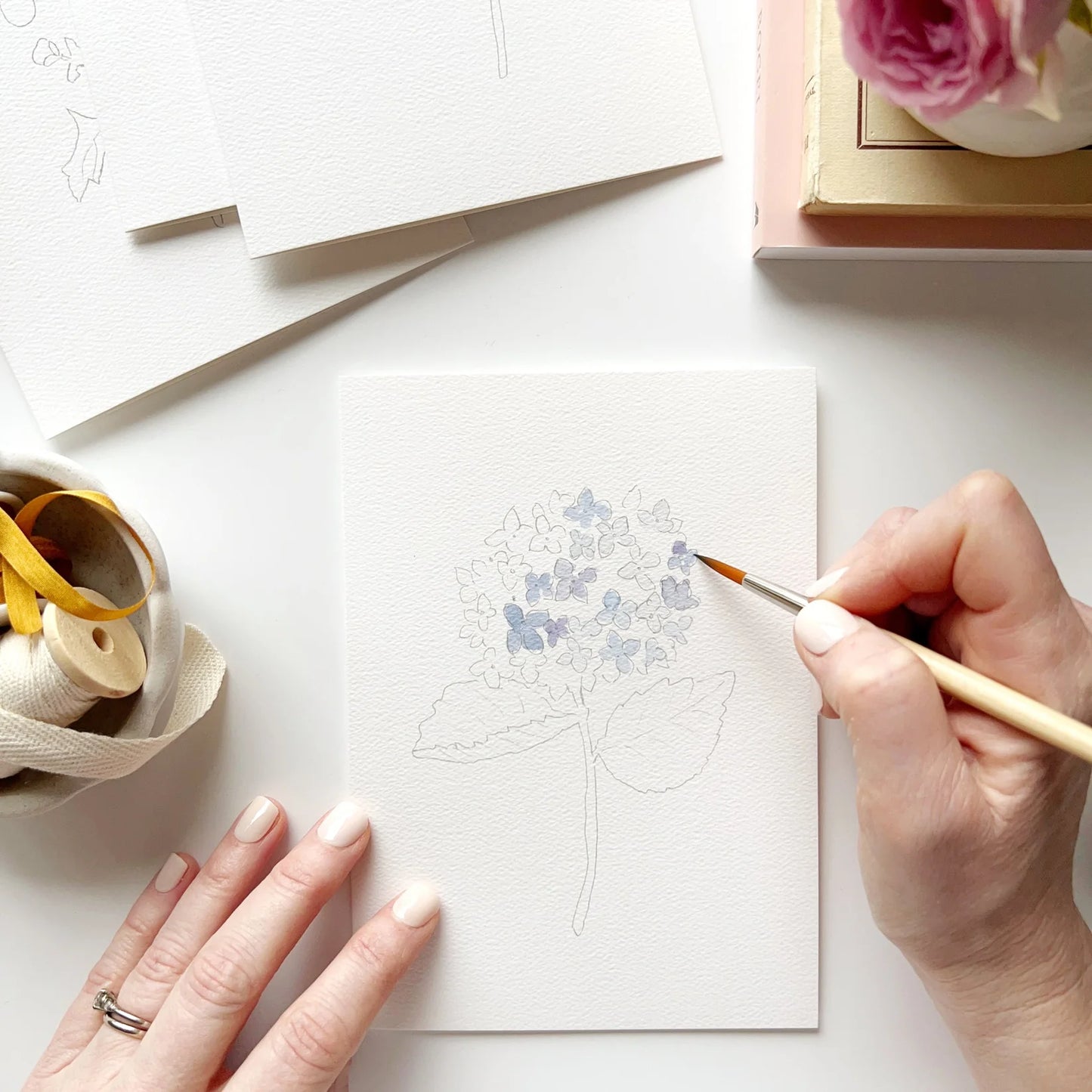 Person painting on a white card with a pen and inkwell on a white surface