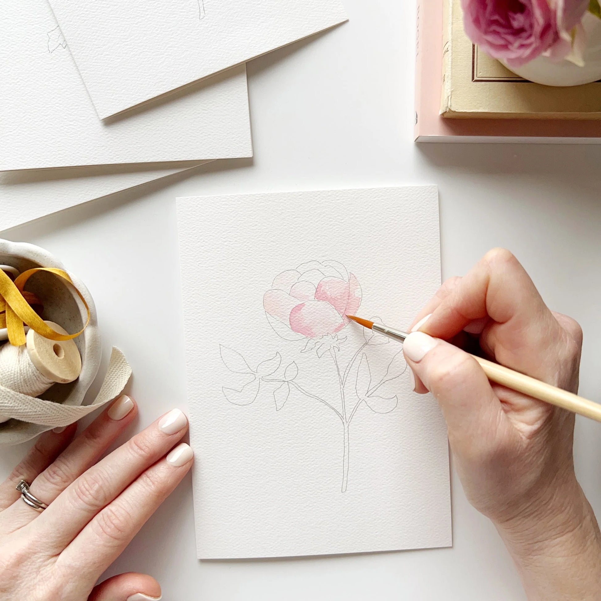 Person painting a pink flower on a white card with a white background