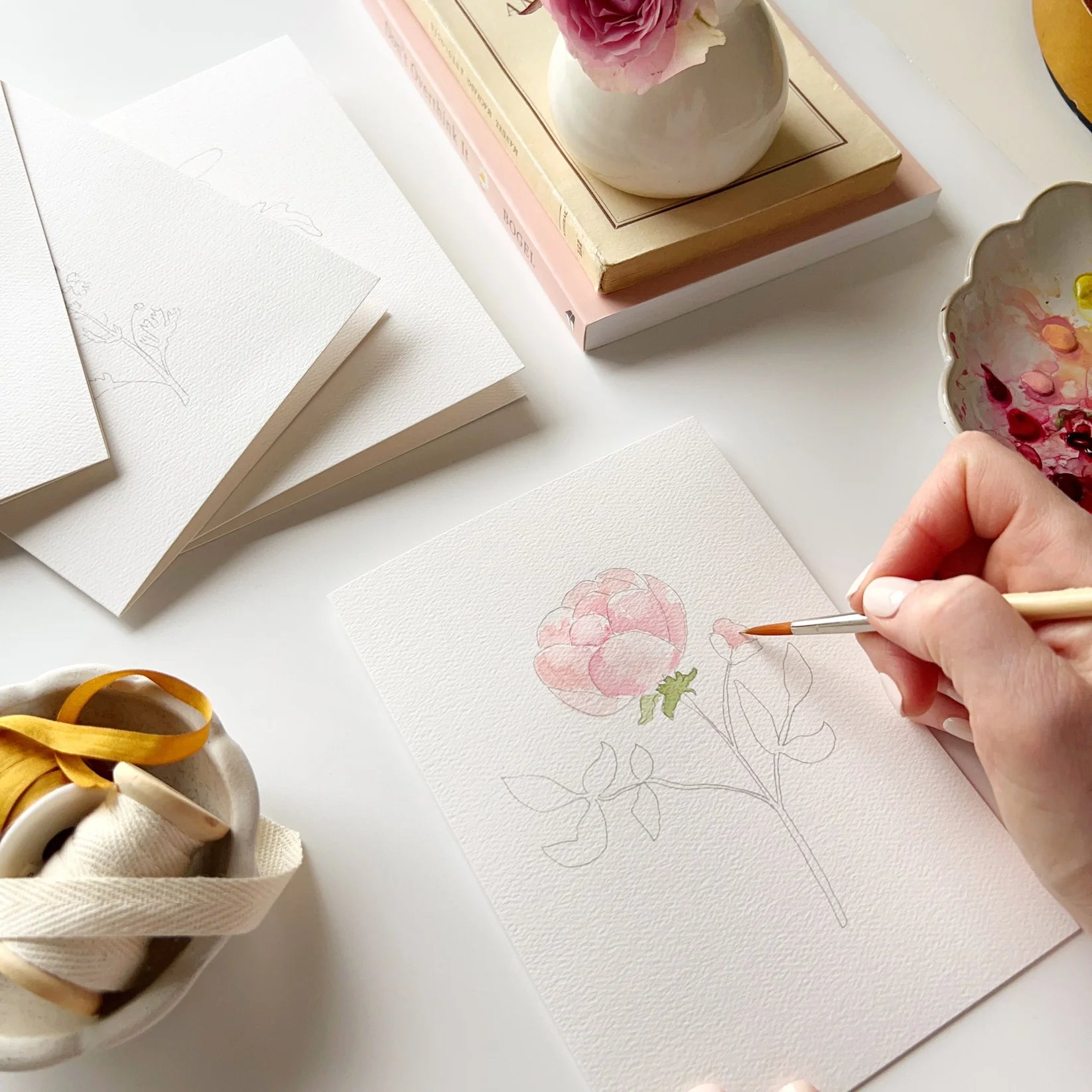 Person painting a pink flower on a white surface with art supplies around