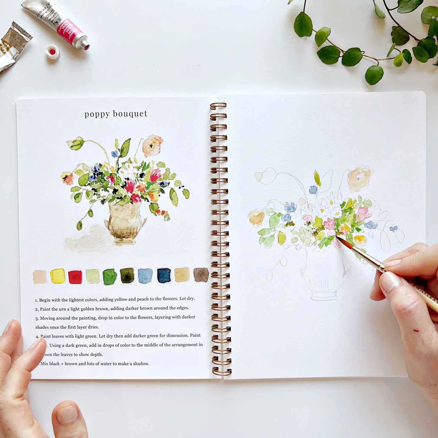 Open book with poppy bouquet floral illustrations and color swatches, hands holding a paintbrush.