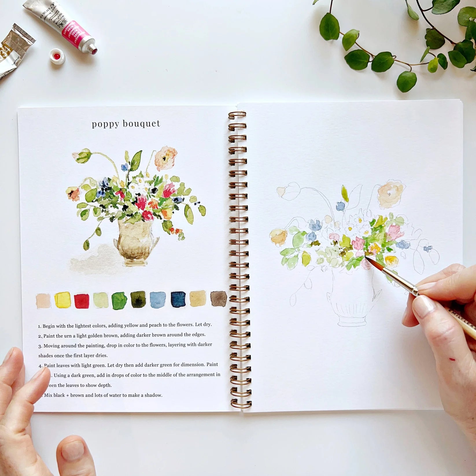 Open book with poppy bouquet floral illustrations and color swatches, hands holding a paintbrush.