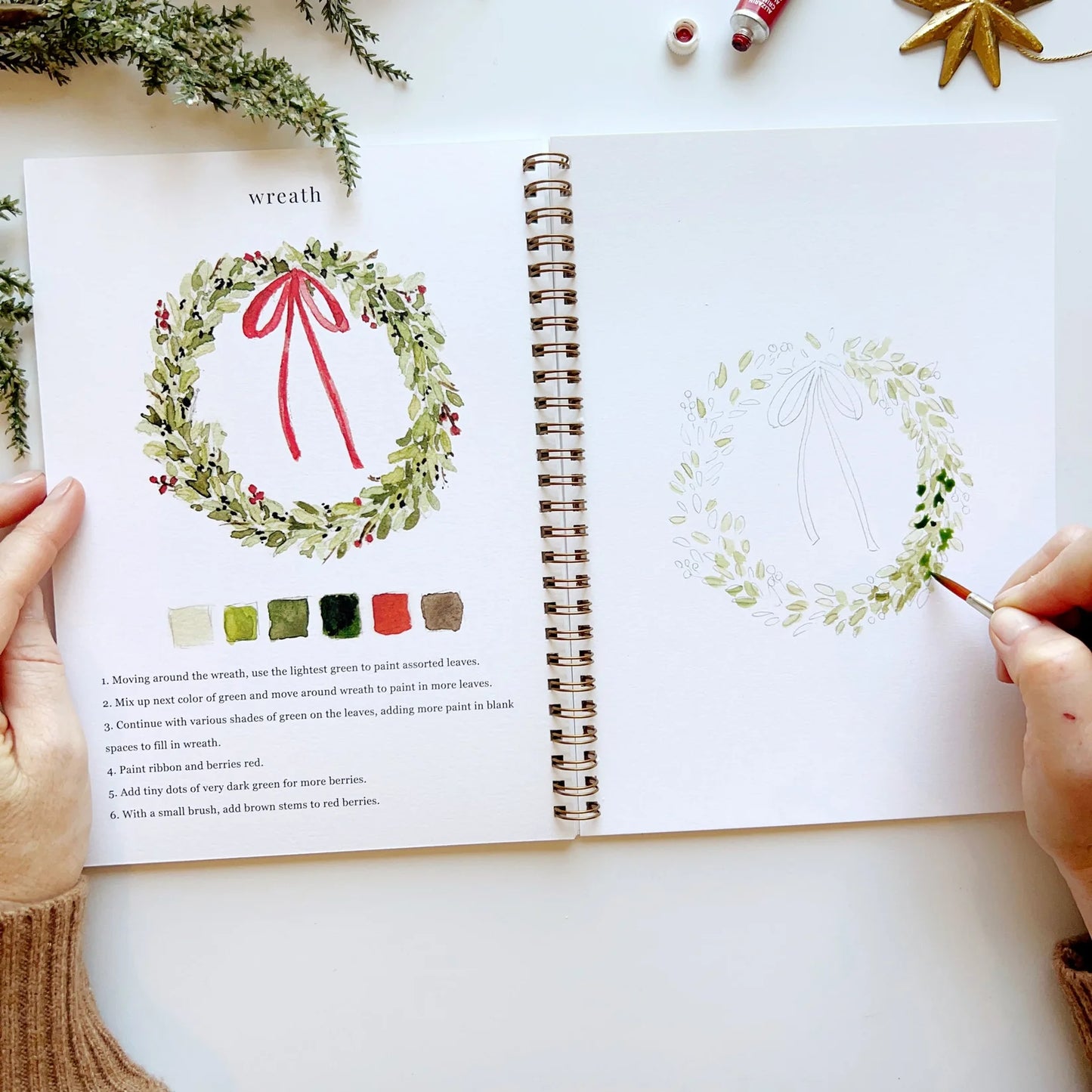 Open book with a wreath illustration and color swatches, hands holding the book.