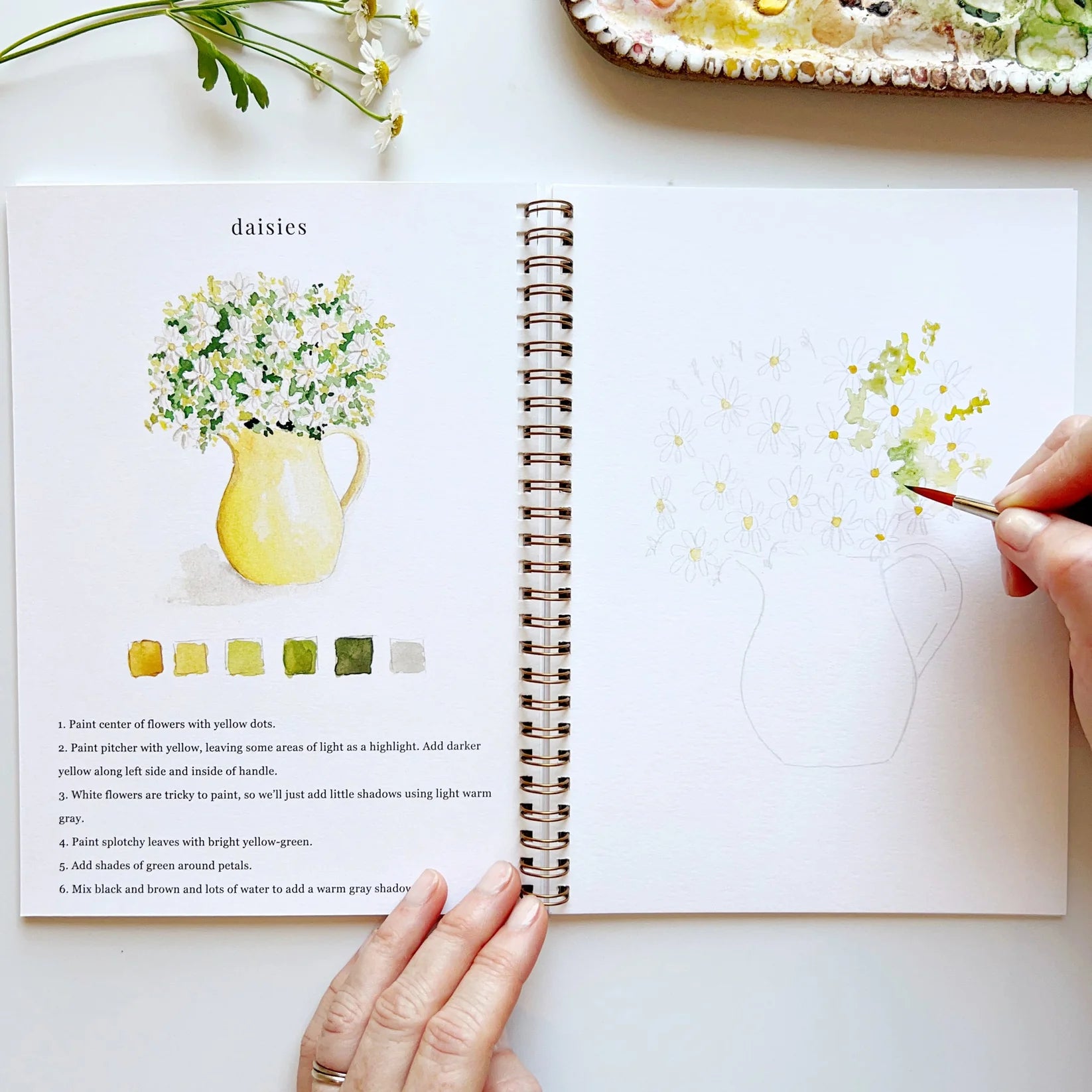 Open book with floral illustrations and a hand holding a paintbrush, with a partially painted dish in the background.