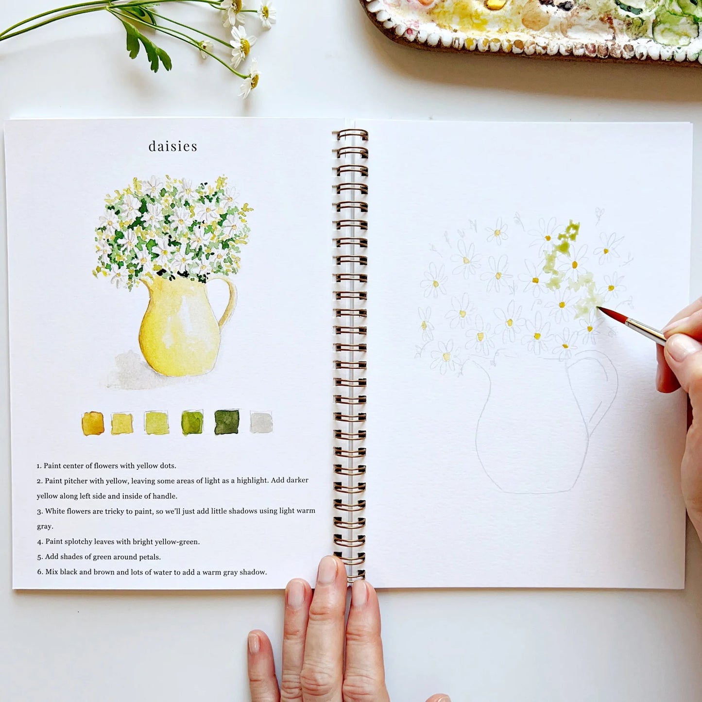 Person painting a flower illustration in a notebook with daisies and a pitcher.