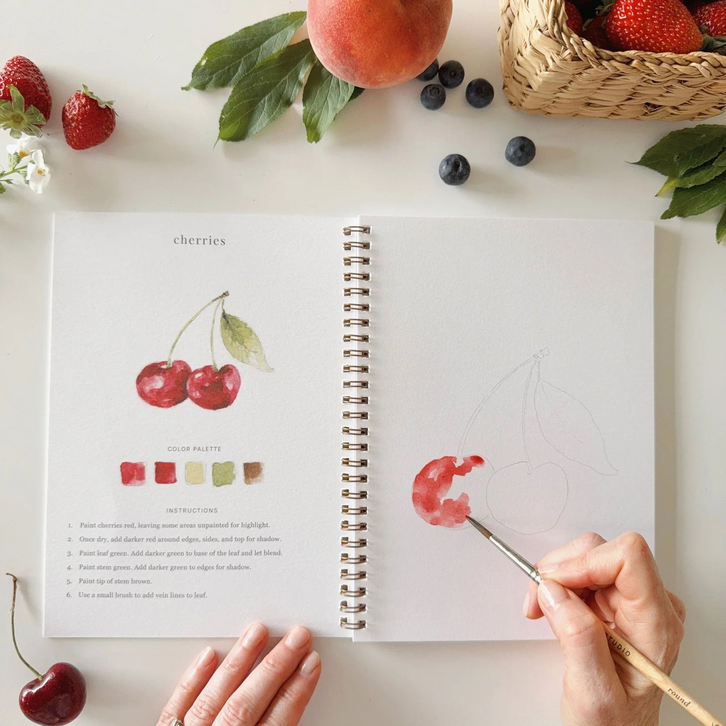 Open notebook with illustration of cherries and a hand holding a paintbrush, surrounded by fruits on a white surface.