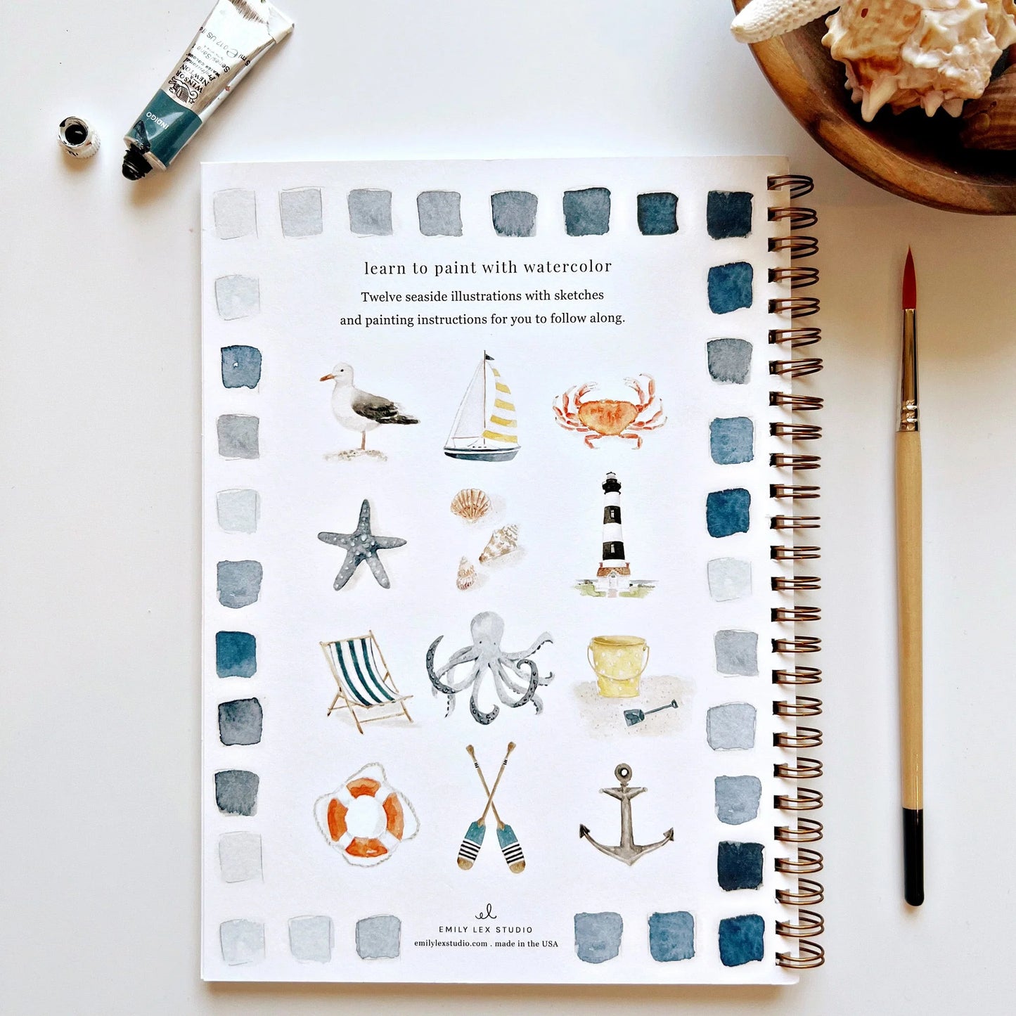 Spiral-bound notebook with watercolor illustrations on a white surface with art supplies.