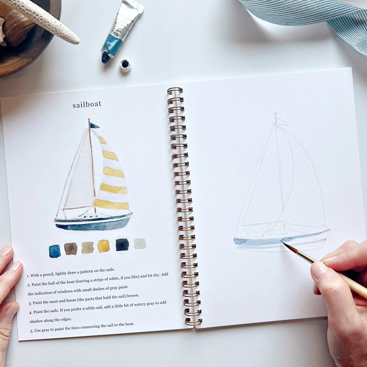 Person drawing a sailboat in a sketchbook with a color chart next to it.