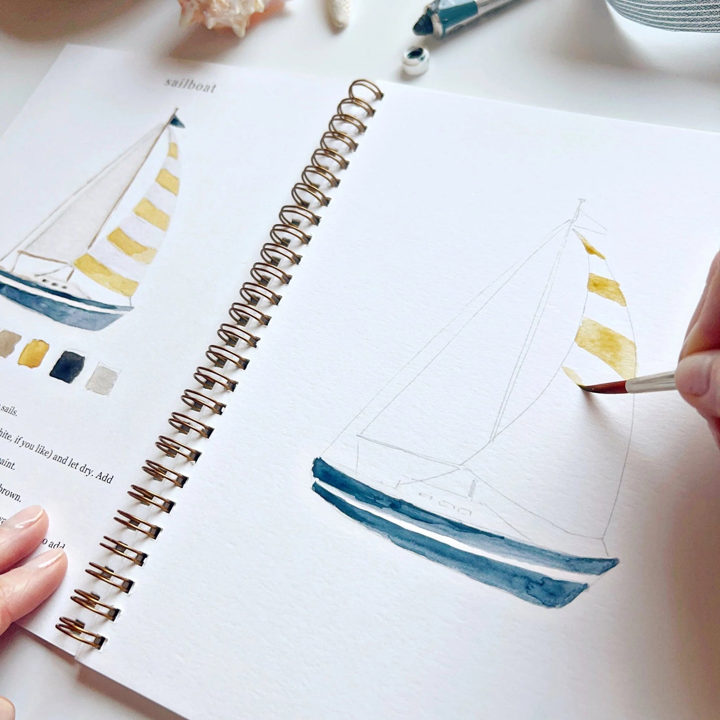 Person painting a sailboat illustration in a sketchbook with watercolors.