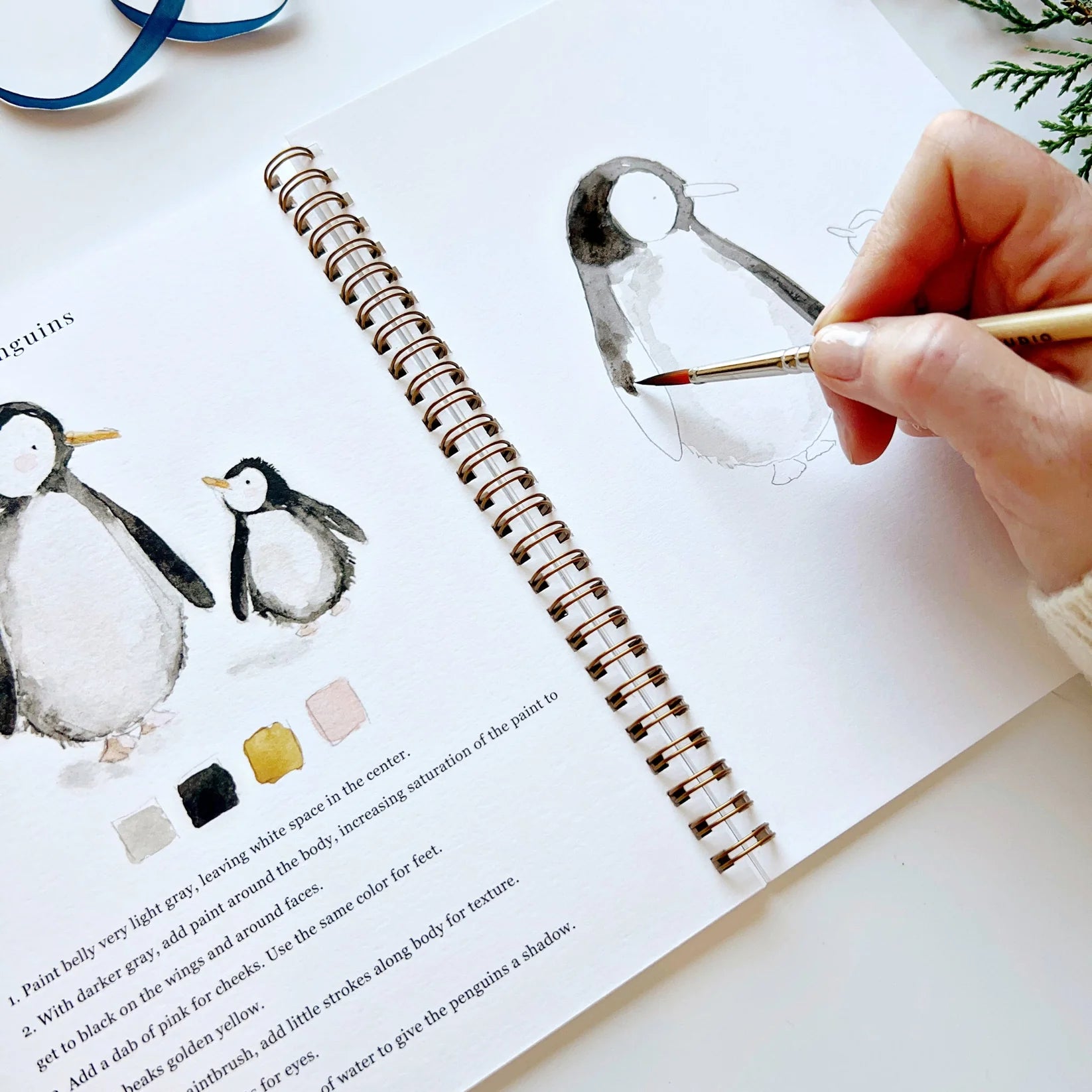 Person painting a penguin illustration with watercolors on a book open to a page with penguin illustrations and color swatches.