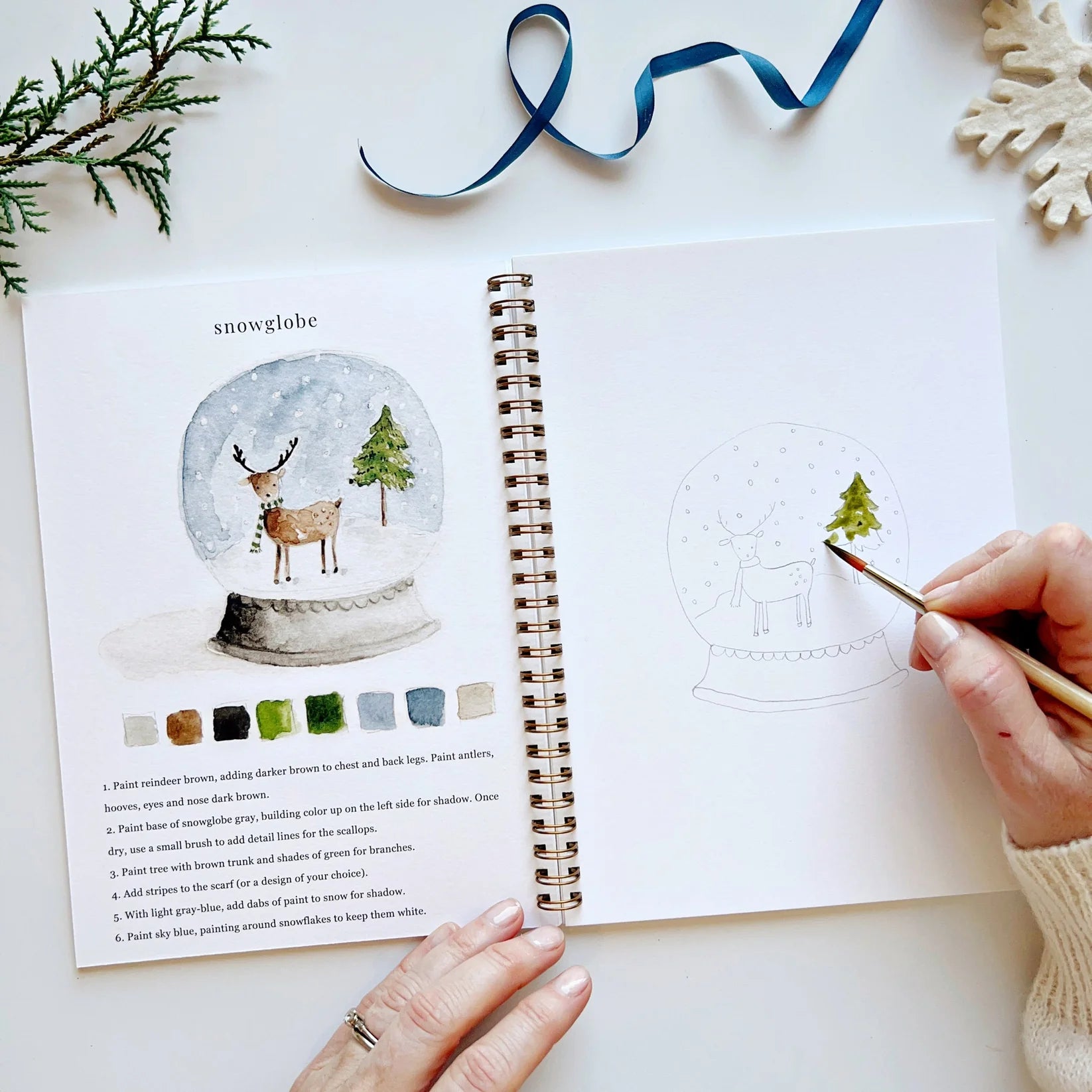 Person painting a snow globe illustration in a notebook with a paintbrush.