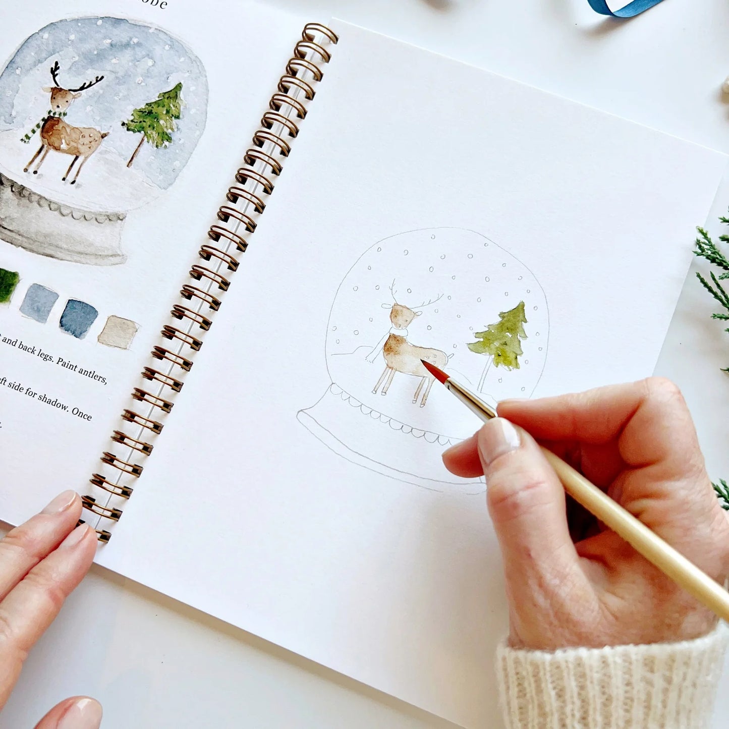 Person painting a snow globe scene with a deer and tree on a notebook