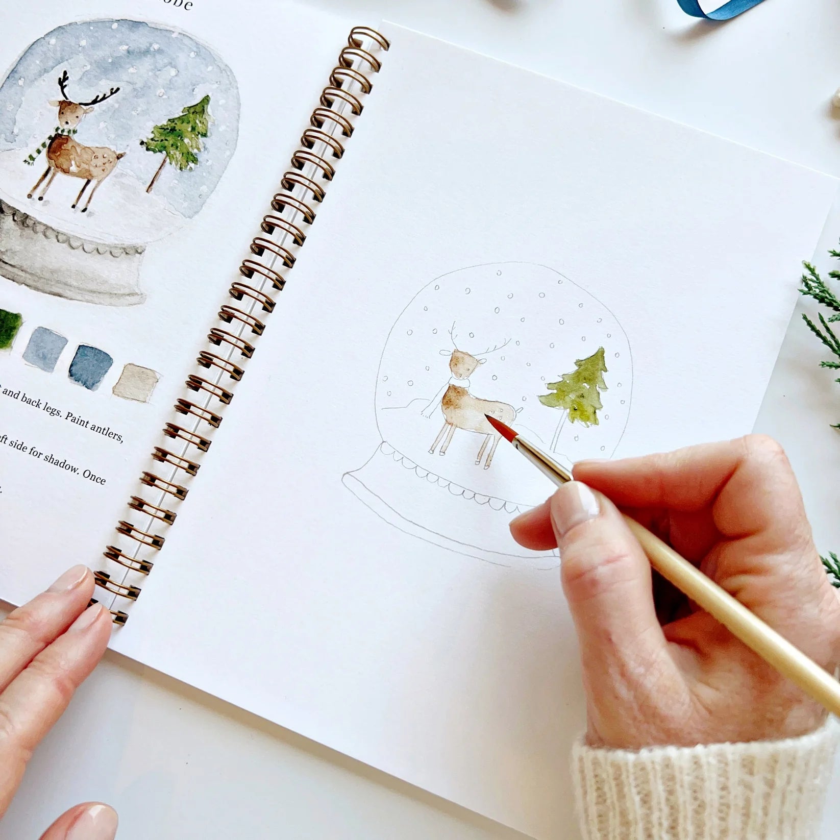 Person painting a snow globe scene with a deer and tree on a notebook